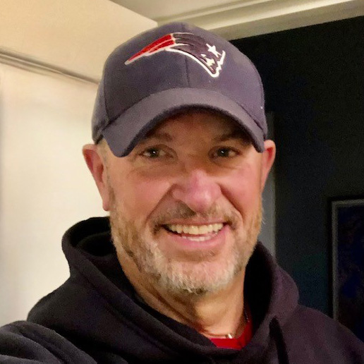 A smiling man with a beard wearing a navy blue New England Patriots baseball cap and a black hoodie, standing indoors.