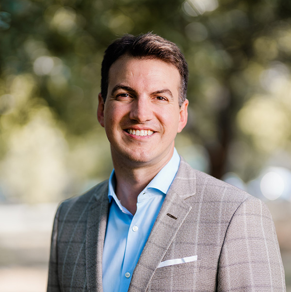 A man in a light gray plaid suit and blue dress shirt smiles outdoors with blurred green trees in the background.